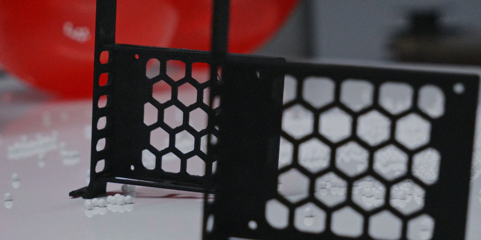 Close-up of two black honeycomb-lattice parts 3D-printed in ESD-safe filament beside scattered foam beads