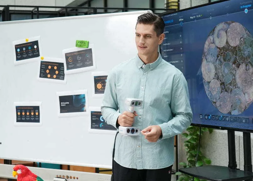 Operator demonstrating the EinScan H2 handheld 3D scanner in a studio with live scan results on a wall display
