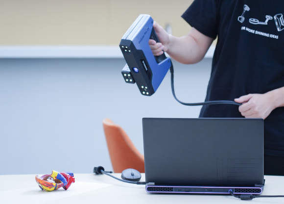 Operator holding the EinScan Pro 2X V2 handheld scanner at a desk while capturing a small colorful part beside a laptop