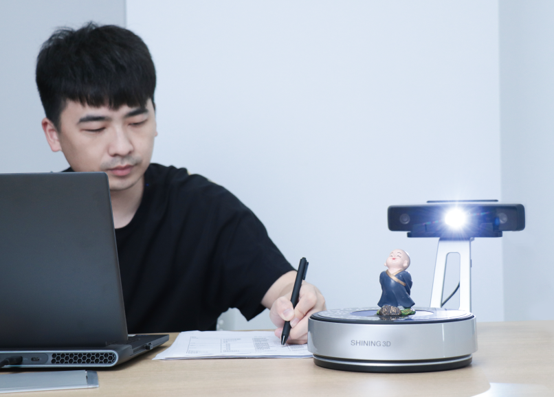 Operator scanning a small figurine on the EinScan SP V2 Auto-Scan turntable beside a laptop running EXScan S