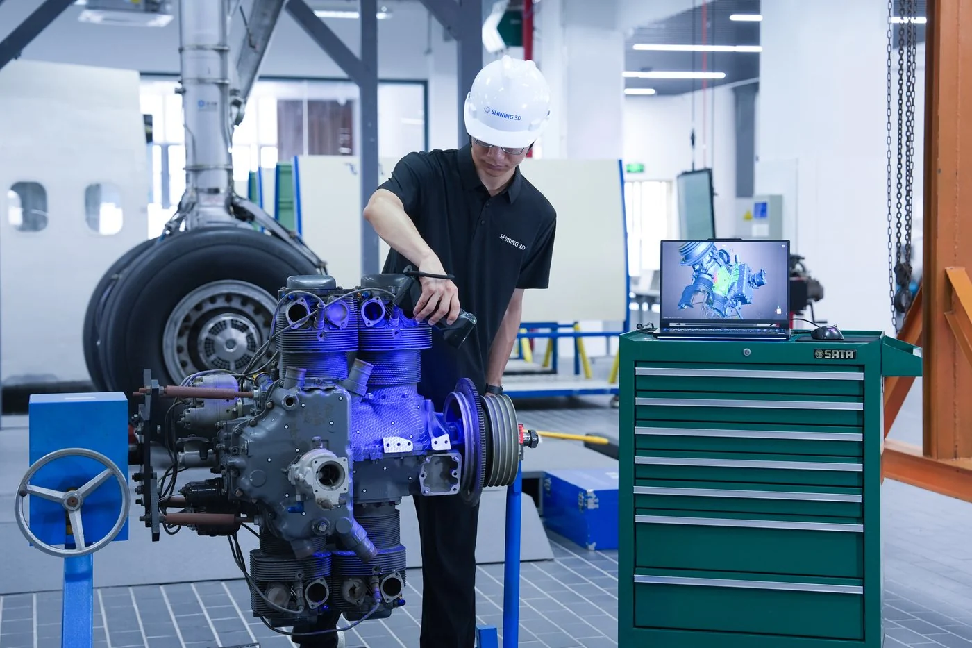 Technician in a hard hat scanning an aircraft piston engine on a stand beside a tool cart and laptop