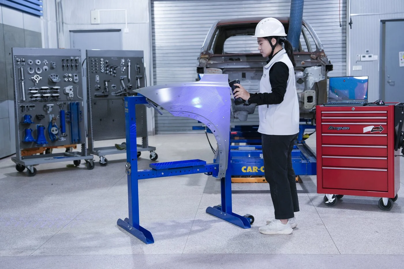 Technician in a hard hat scanning a car fender held in a blue fixture on the shop floor